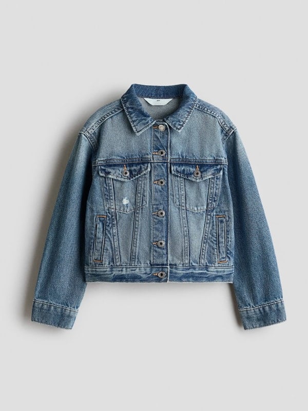 Mid-wash cropped denim trucker jacket with chest flap pockets and silver buttons, displayed flat on a light grey background.