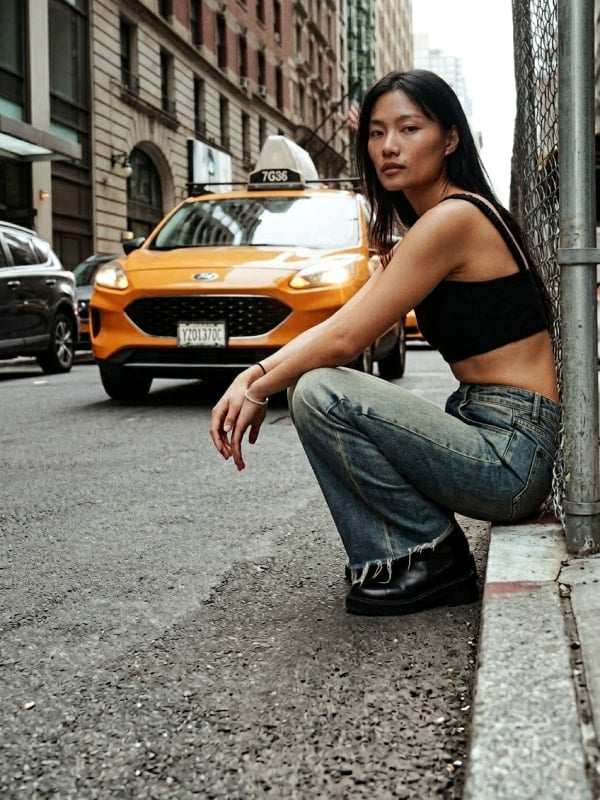 Asian woman crouched on a New York City sidewalk in a black square-neck tank top, baggy faded jeans, and black ankle boots, with a yellow taxi pulling up behind her.
