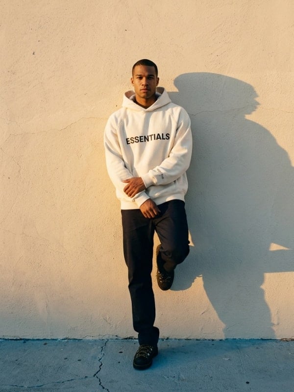 Man in a cream Essentials hoodie and black jeans leaning against a stucco wall in warm afternoon sunlight, casting a long shadow.