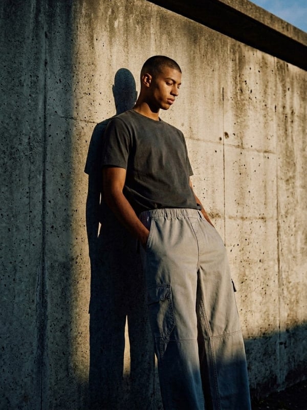 Man in a charcoal tee and light grey washed cargo pants leaning against a sunlit concrete wall in golden hour light.