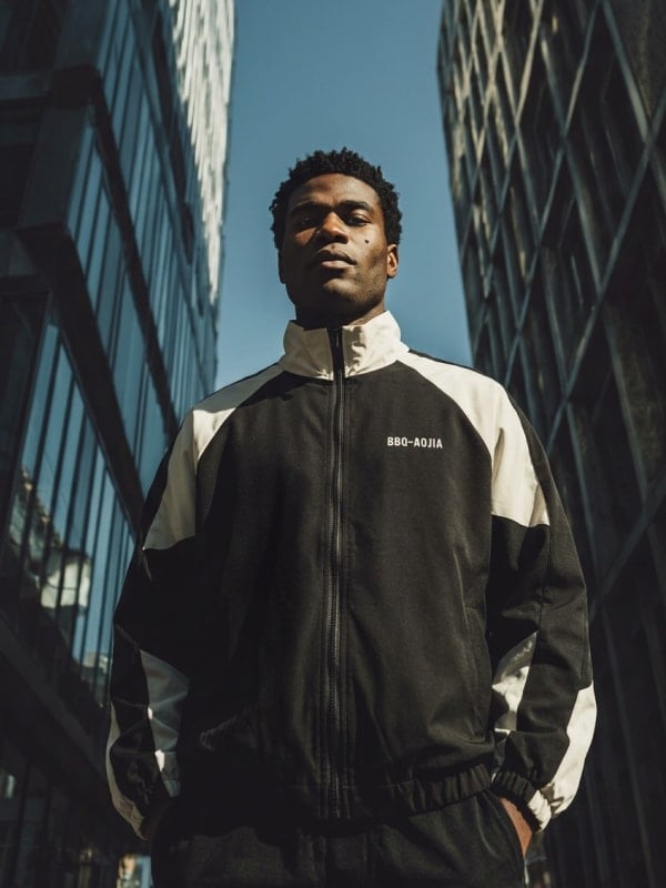 Low-angle shot of a man in a black and cream retro track jacket standing between mirrored glass office towers under a bright blue sky.