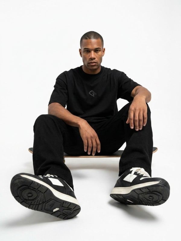 Man seated on a black skateboard in a clean white studio, wearing an oversized black tee, black trousers, and chunky black-and-white sneakers.