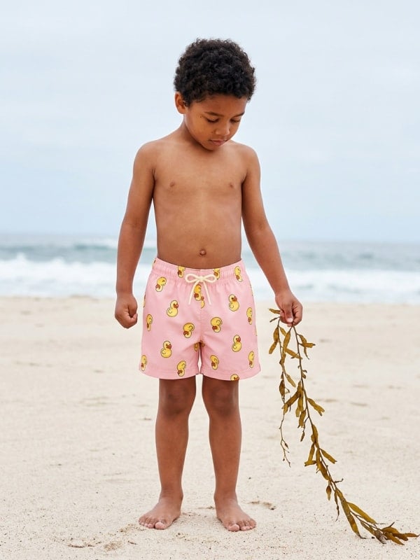 Young boy in pink rubber-duck-print swim trunks standing on a sandy beach holding a strand of seaweed, with the ocean behind him.