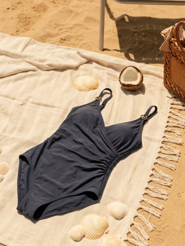 Navy blue one-piece swimsuit laid on a striped beige beach blanket with seashells, a halved coconut, and a woven rattan bag in dappled sunlight.